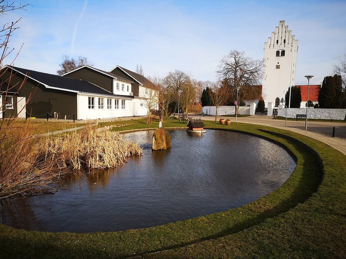 A village pond beside houses and a white church tower in Denmark.